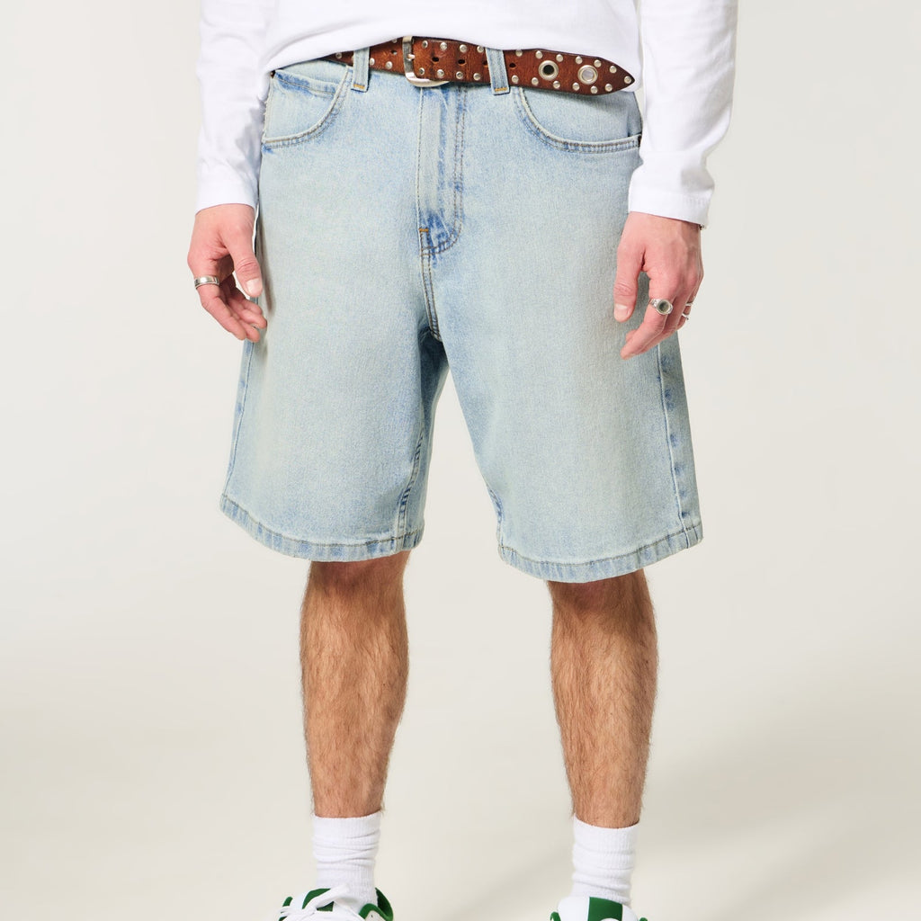 A person wearing light-wash denim shorts with a brown patterned belt, white long-sleeve shirt, white socks, and white sneakers with green accents, shown from the waist down on a plain light beige studio background.