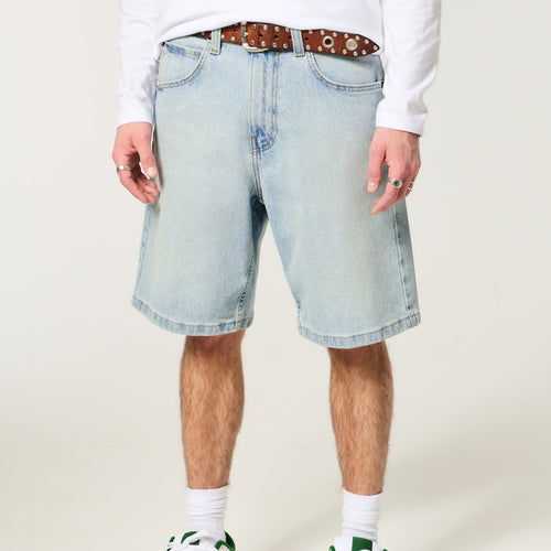 A person wearing light-wash denim shorts with a brown patterned belt, white long-sleeve shirt, white socks, and white sneakers with green accents, shown from the waist down on a plain light beige studio background.