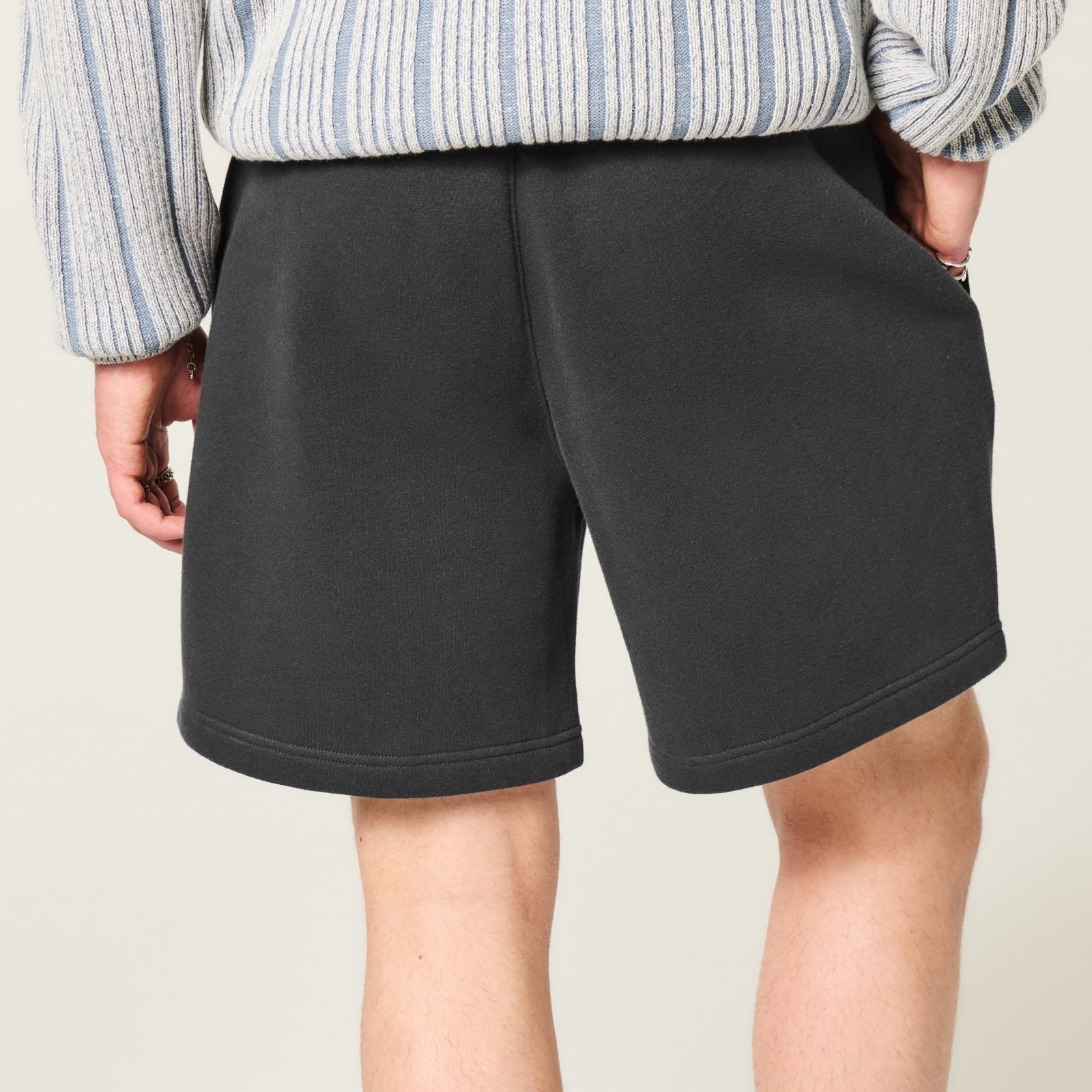 A person shown from the back wearing dark charcoal gray shorts and a light blue and white striped long-sleeve top, against a plain light beige background.